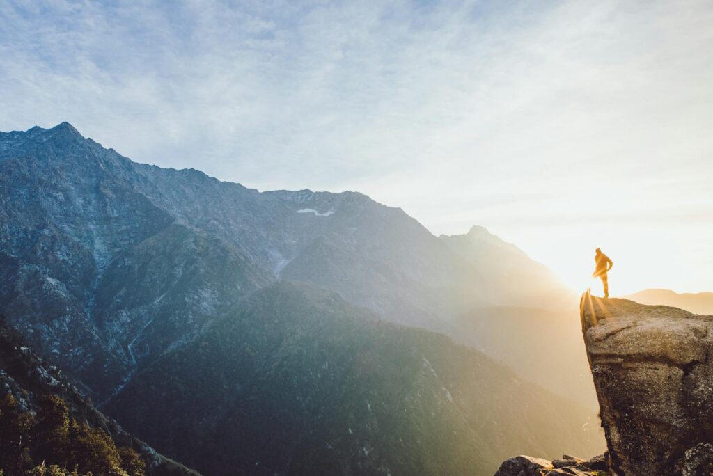 A lone hiker stands on a cliff, overlooking a mountain range at sunrise in India.